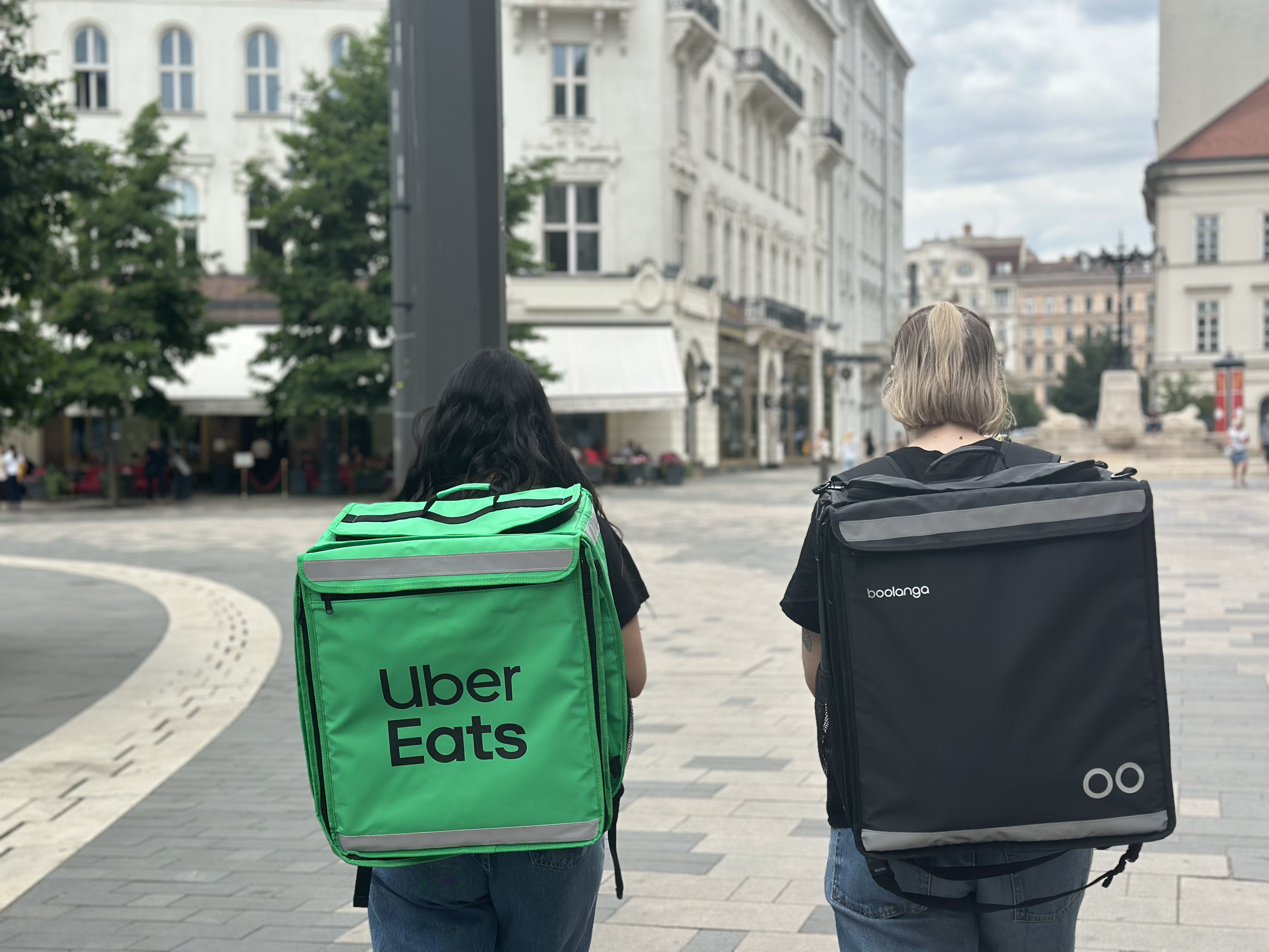 Two delivery riders walking down a Budapest street, one carrying an Uber Eats bag and one carrying a Boolanga bag